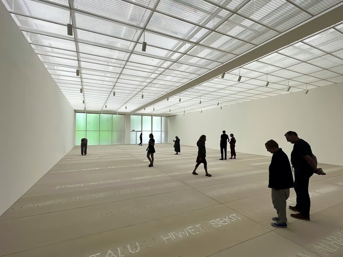 Persone osservano un'installazione artistica minimalista con nomi sul pavimento di una galleria.